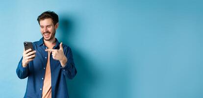 Image of handsome smiling man reading mobile screen and pointing at phone, checking out online promo or smartphone app, standing on blue background photo
