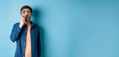 Impressed man look in awe through magnifying glass, drop jaw and stare at camera, standing on blue background photo