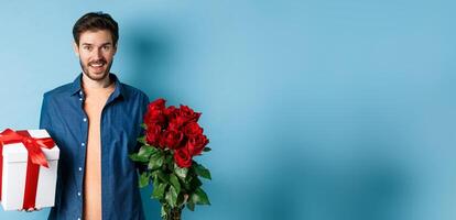 Love and relationship concept. Happy young man bring flowers and gift on romantic date. Boyfriend wish bouquet of roses and present standing on blue background photo