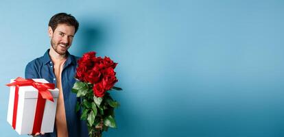Love and Valentines day concept. Happy guy winking and smiling at camera, presenting a gift and bouquet of flowers to girlfriend, standing over blue background photo