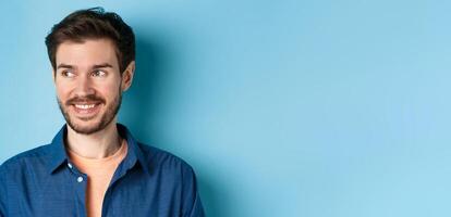 Close up of handsome confident man with beard, looking at empty space and smiling, standing on blue background photo