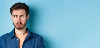 Miserable and disappointed guy acting childish, sulking and looking left at empty space with offended face, standing on blue background photo