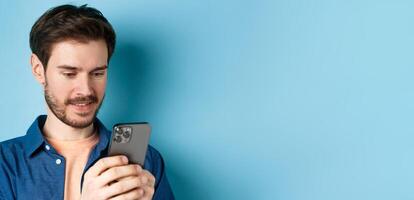 Close up portrait of happy young man reading mobile sreen, networking in smartphone, standing on blue background photo