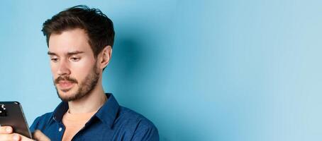 Close up of handsome bearded man reading mobile phone message, looking at screen serious, standing on blue background photo