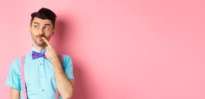 Image of handsome funny man in bow-tie looking thoughtful left and touching lip, thinking what to choose, making decision, standing over pink background photo