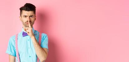 Angry and grumpy man in funny bow-tie shushing at camera, frowning and demand silence, tell to be quiet, scolding someone loud, standing on pink background photo