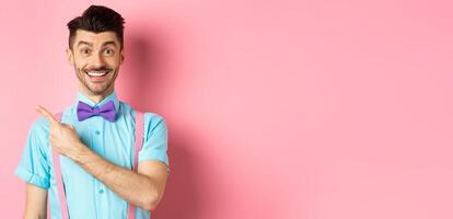 Portrait of handsome happy man showing logo, pointing left and smiling at camera, recommending shop offer, standing on pink background photo