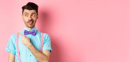 Funny man express disappointment, pointing at something bad and showing tongue, looking left at promo, standing on pink background photo