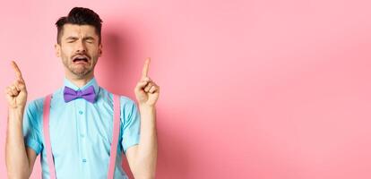 Sad and miserable guy sobbing and crying, pointing fingers up and something disappointed, standing upset on pink background photo