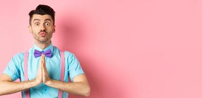 Young guy with cute face pucker lips and asking please, begging for favour, need something and lookin gpleading at camera, standing over pink background photo