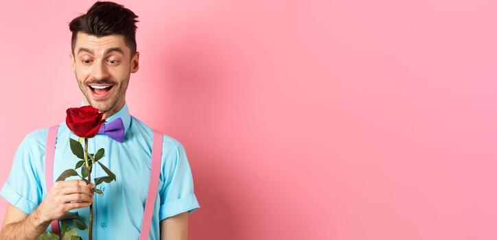 san valentin día y romance concepto. contento novio mirando a rojo Rosa mientras esperando para amante en corbata de moño, en pie en rosado romántico antecedentes foto