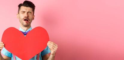 Valentines day concept. Sad and lonely man feeling heartbroken, being rejected, showing big red heart cutout and crying from break-up, standing on pink background photo