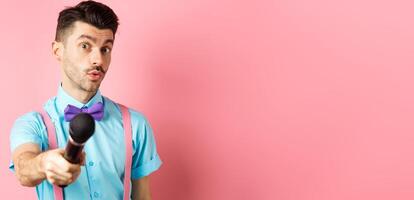 Handsome male show host stretch out hand with microphone, asking for interview or comments, standing on pink background photo