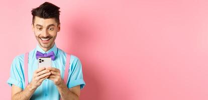 Online shopping. Happy guy looking at smartphone screen, reading message and smiling, standing on pink background photo