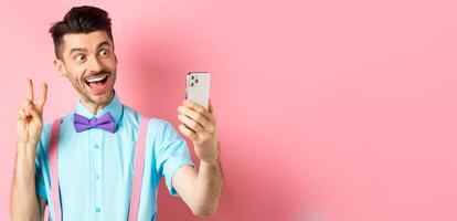Technology concept. Funny man with moustache and bow-tie taking selfie on smartphone, showing peace sign and smiling at mobile camera, pink background photo
