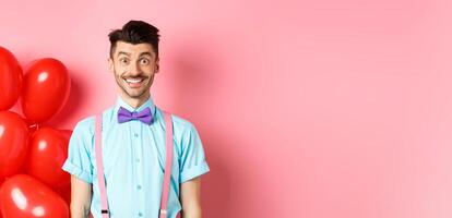 Valentines day concept. Image of handsome young man looking excited and surprised, smiling while standing on romantic pink background near heart balloons photo