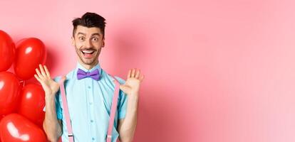 Valentines day concept. Happy young man looking surprised, raising hands up joyful, celebrating near big red hearts and pink background photo