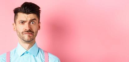 Close-up of anxious young guy with moustache, biting lip and frowning, looking worried over small mistake, standing on pink background photo