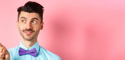 Close-up of caucasian male model with moustache and bow-tie, looking and pointing left with intrigued smile, showing promo offer, standing in bow-tie on pink background photo