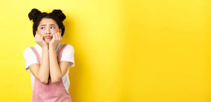 Sad moody asian girl, lean face on hands, looking left with sulking expression, standing in summer clothes on yellow background photo