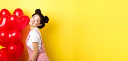Valentines day concept. Stylish teenage asian girl posing near red hearts balloons, wear outfit for romantic date, standing happy on yellow background photo
