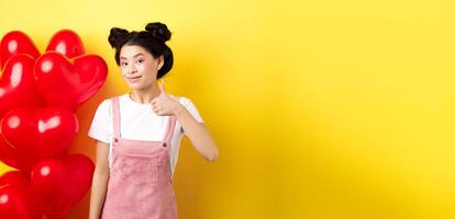 Fashionble asian woman in romantic outfit with make-up, showing thumb up and smiling, praising Valentines day offer, standing near red heart balloons, yellow background photo