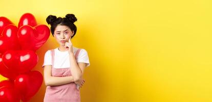 Valentines day concept. Unamused and bored asian girlfriend staring skeptical at camera, waiting for something near red hearts balloons, yellow background photo