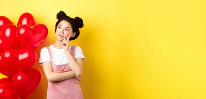 Valentines day concept. Pensive asian woman looking left, thinking about romantic date, standing near red hearts balloons on yellow background photo