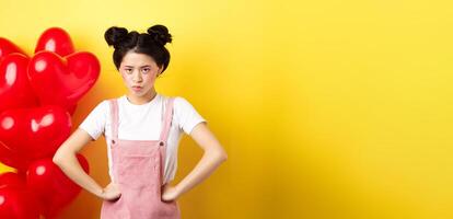 Valentines day and relationship concept. Disappointed asian girlfriend sulking, looking offended at camera, mad at boyfriend, standing near heart balloons, yellow background photo