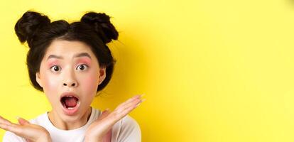 Close-up of excited asian woman with glamour makeup and hairstyle, screaming of amazement, open mouth and look at camera in awe, yellow background photo
