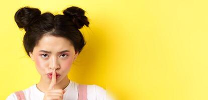 Close up portrait of angry asian girl shushing, press fingers on lips and tell be quiet, scolding loud person, need silence, telling big secret, standing on yellow background photo