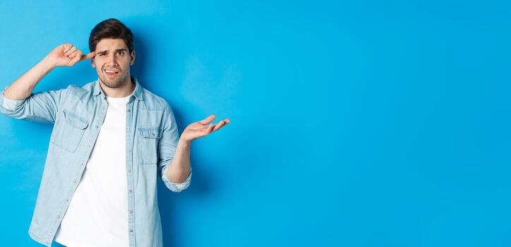 confuso hombre regaño persona para siendo estúpido, señalando dedo a cabeza y encogiéndose de hombros, mirando perplejo, desaprobar comportamiento, en pie en contra azul antecedentes foto