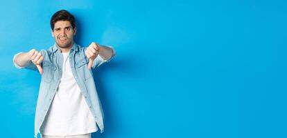 Disappointed man showing thumbs down and grimacing at something awful, dislike product, judging bad advertisement, standing against blue background photo