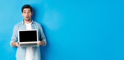 Man showing advertisement on laptop screen and looking amazed, saying wow and looking at camera, standing against blue background photo
