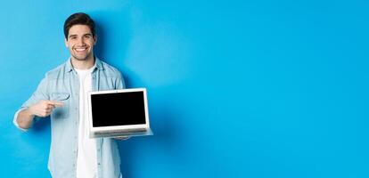 Handsome young man pointing finger at screen of computer, smiling pleased, showing promo in internet or website, standing over blue background photo