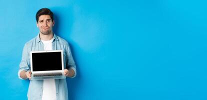 Skeptical and displeased bearded guy showing laptop screen and grimacing, having doubts, standing over blue background in casual clothes photo