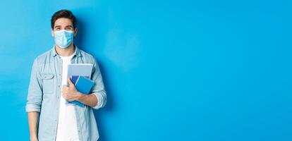 Education, covid-19 and social distancing. Guy student in medical mask looking happy, holding notebooks, standing over blue background photo