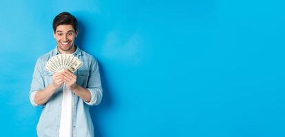 Excited successful man counting money, looking satisfied at cash and smiling, standing over blue background photo