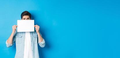 Shy and nervous guy hiding face behind blank piece of paper, peeking with worried look, showing your logo, standing over blue background photo