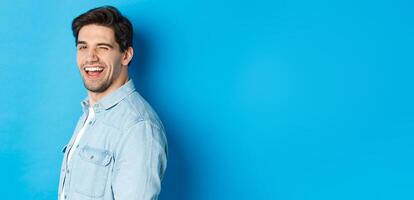 Happy and confident man turn head at camera, winking and smiling, standing over blue background photo