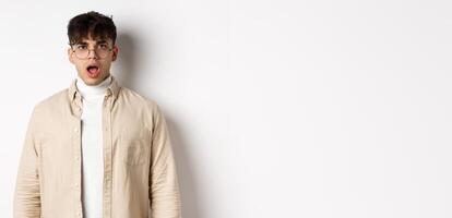 Portrait of shocked young man in glasses drop jaw, open mouth and stare amazed at camera, looking at something impressive, standing on white background photo