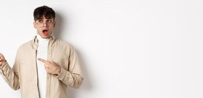 Amazed young man in glasses drop jaw, saying wow, pointing and looking left at awesome promo offer, stare with disbelief and impressed face, white background photo