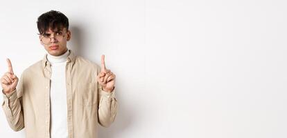 Portrait of young male model in glasses looking suspicious and frowning, pointing fingers up with disbelief, standing on white background photo