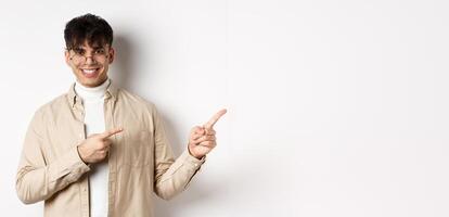 Handsome caucasian man in glasses smiling, pointing fingers at upper right corner, showing logo on white background photo