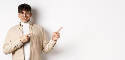 Proud and happy young man in glasses showing logo, pointing fingers at upper right corner and smiling, standing on white background photo