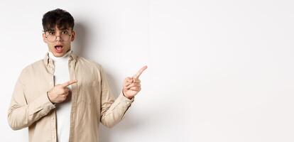 Shocked young man in glasses pointing fingers right at empty space, asking about advertisement, standing on white background photo