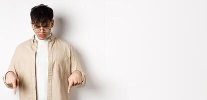 Confused frowning guy in glasses looking, pointing fingers down at something strange, standing puzzled on white background photo