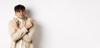 Indecisive funny young man having troubles with making choice, pointing fingers sideways and frowning perplexed, deciding between two variants, standing on white background photo