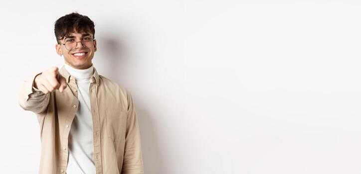 Handsome modern guy in glasses pointing at camera, smiling and choosing you, recruiting or inviting to event, standing on white background photo