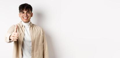 Image of happy young man showing thumb up and smiling satisfied, praise good job, say well done and look pleased, standing on white background photo